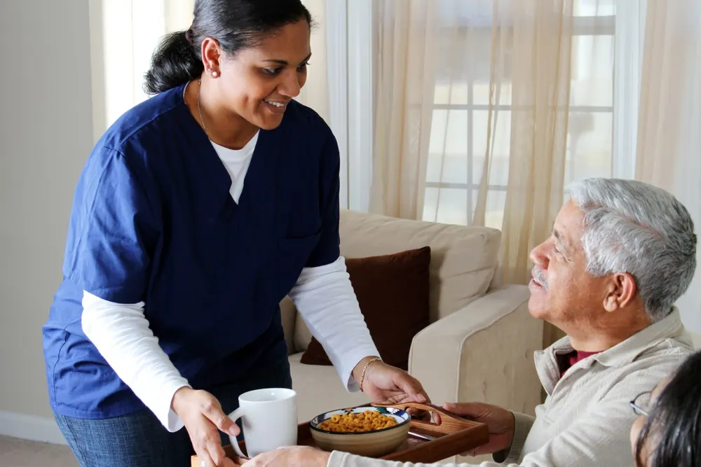 A caregiver serves a meal to an older adult in a living room.